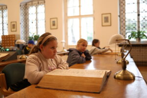 Wnętrze biblioteki z kilkoma osobami siedzącymi przy długim drewnianym stole. Na pierwszym planie osoba czyta dużą otwartą książkę. W tle inne osoby również zajęte są czytaniem lub pisaniem.