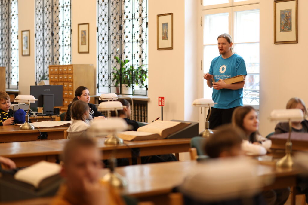 Grupa osób siedzi przy stołach w bibliotece. Na pierwszym planie leżą otwarte książki, a w głębi osoba w niebieskiej koszulce trzyma notatnik i mówi do grupy.