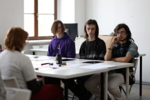 Cztery osoby siedzące przy stole w jasnym pomieszczeniu. Trzy osoby po jednej stronie stołu, jedna po drugiej. Na stole leżą dokumenty, długopisy i kubek.