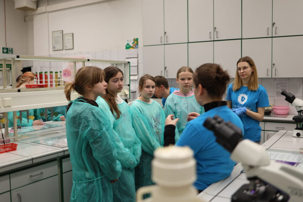 Grupa osób w laboratorium; część z nich nosi zielone fartuchy ochronne