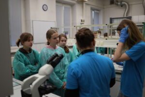 Grupa osób w laboratorium; część z nich nosi zielone fartuchy ochronne